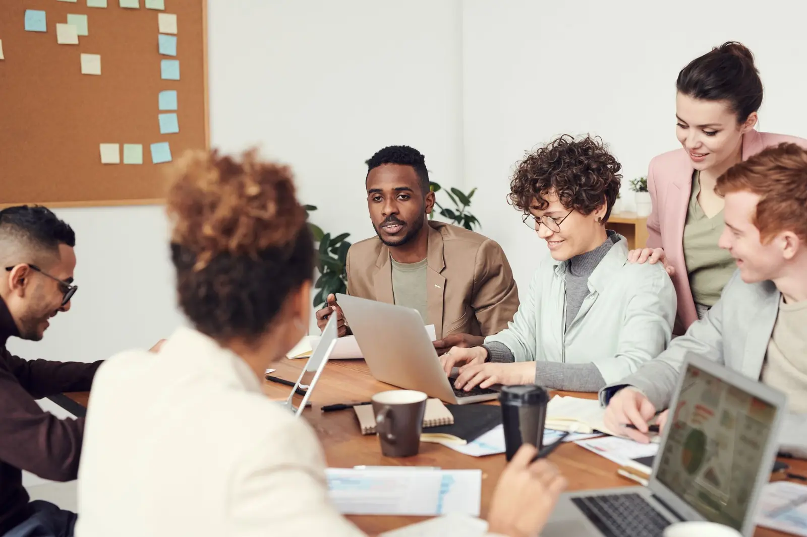 A diverse team gathered around a table with laptops in front of a wall of sticky notes during a planning session